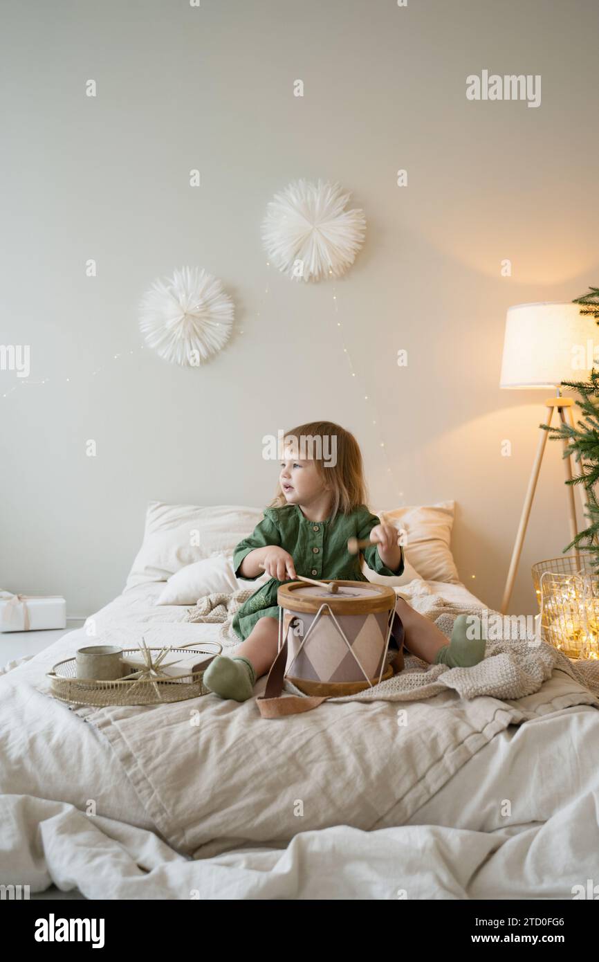 Kleines Kind spielt auf einem gemütlichen Bett Trommeln in einem warm beleuchteten Zimmer mit festlicher Dekoration. Stockfoto