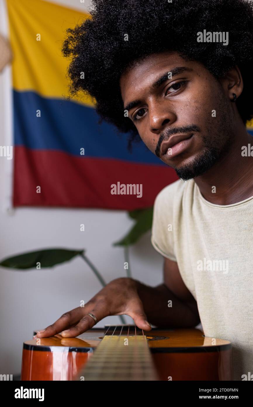 Nahaufnahme eines fokussierten jungen Mannes mit Afro-Haaren, der Akustikgitarre spielt, während er am Wochenende zu Hause gegen kolumbianische Flagge sitzt Stockfoto