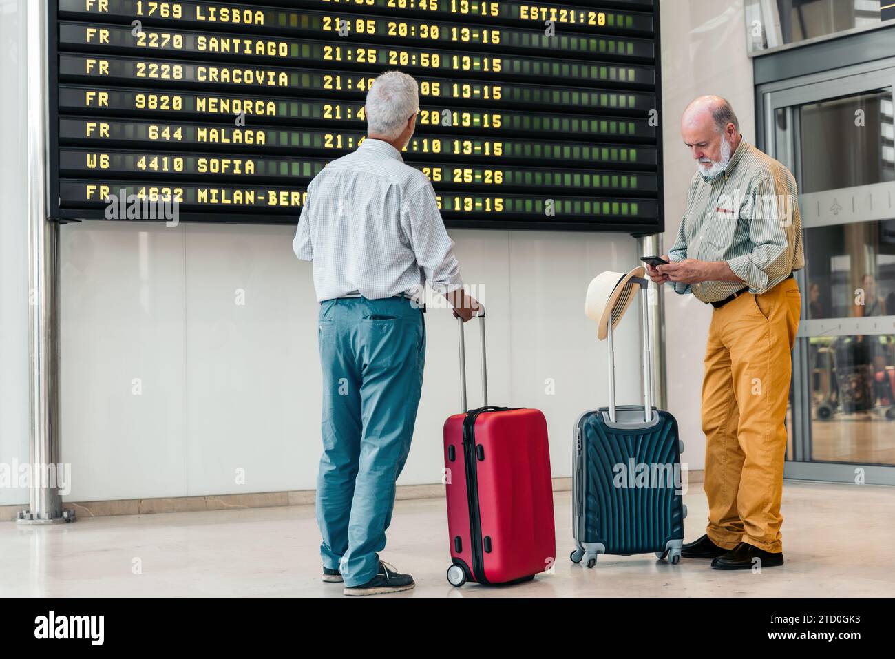 Rückansicht eines anonymen männlichen Seniortouristen in lässiger Kleidung, der die Abflugplantafel am zeitgenössischen Flughafen ansieht, während sein Freund auf seinem schreibt Stockfoto