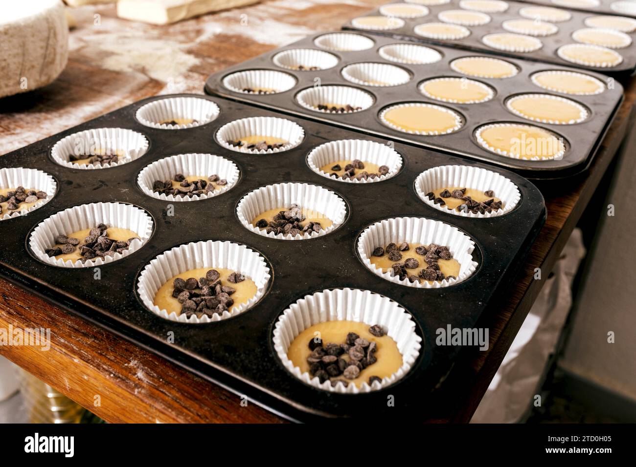 Großaufnahme eines Muffins-Tabletts aus Metall mit Cupcake-Teig und Schokoladenchips in Papierformen, die auf dem Tisch zum Backen im Backhaus angeordnet sind Stockfoto