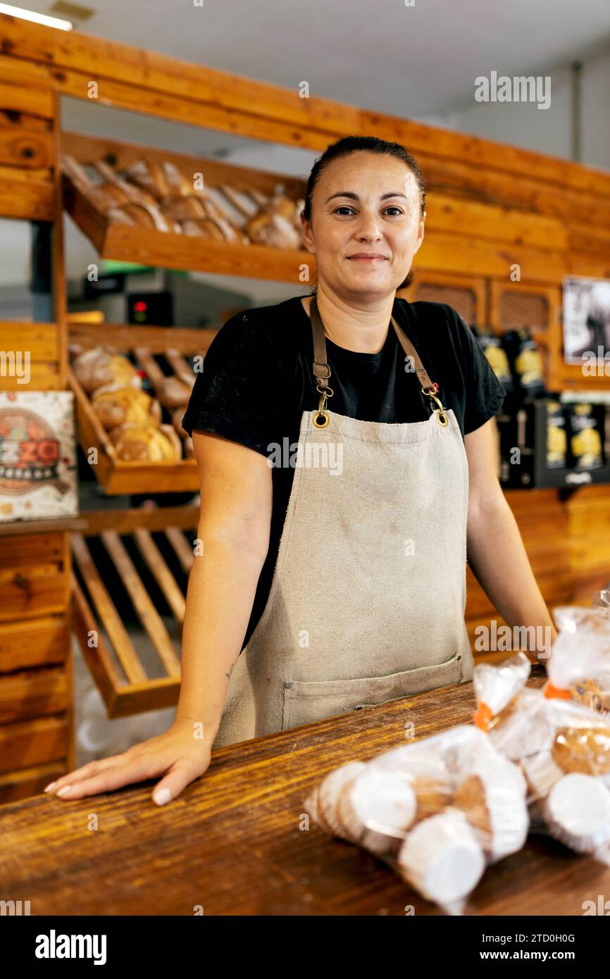Porträt einer selbstbewussten positiven Verkäuferin in Schürze mit Händen auf der Theke, die neben frisch gebackenem Brot auf Holzregalen in Bak steht Stockfoto
