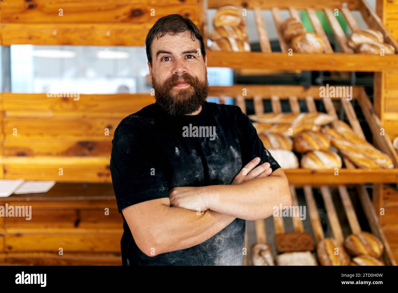 Porträt eines selbstbewussten, positiv bärtigen, Reifen Mannes mit überkreuzten Armen, der vor Holzregalen mit frisch gebackenem Brot in der Bäckerei steht Stockfoto