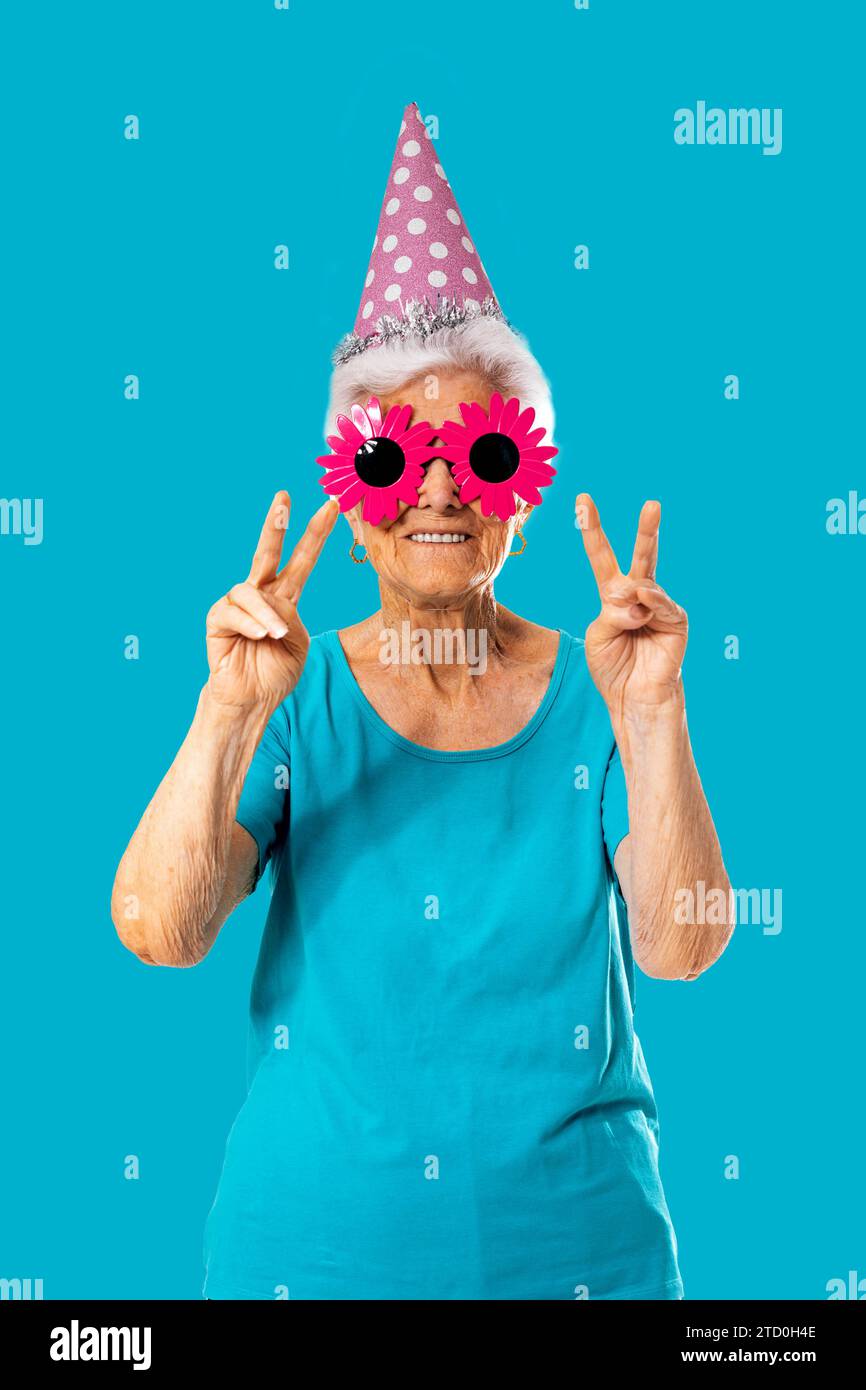 Lächelnde ältere Frau mit lustiger rosafarbener Sonnenbrille und Geburtstagskappe, während sie Friedensgeste vor blauem Hintergrund zeigt Stockfoto