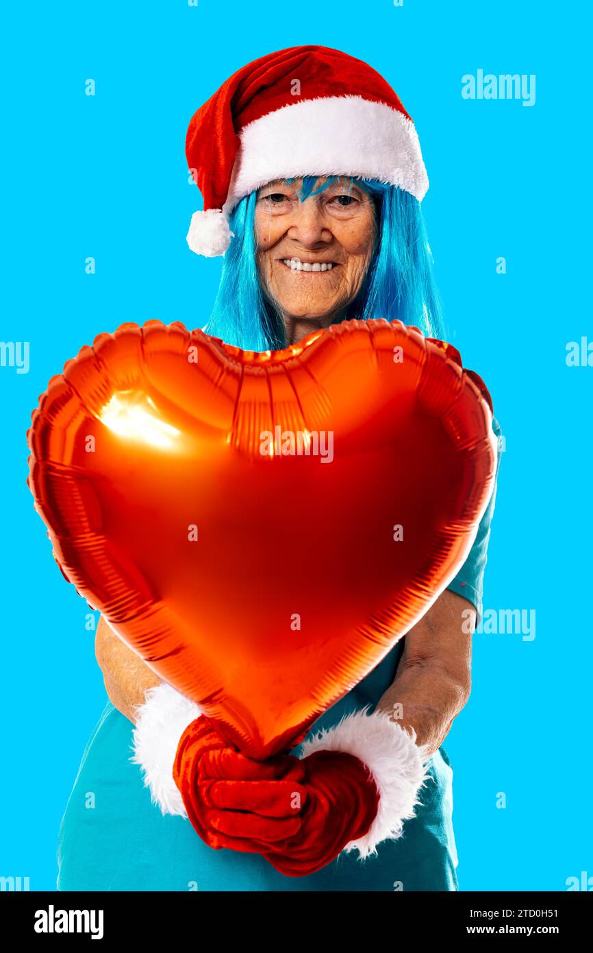 Fröhliche ältere Frau in blauer Perücke, Weihnachtsmannmütze und Handschuhen, die während der Weihnachtsfeier einen roten Herzballon vor blauem Hintergrund demonstrieren Stockfoto