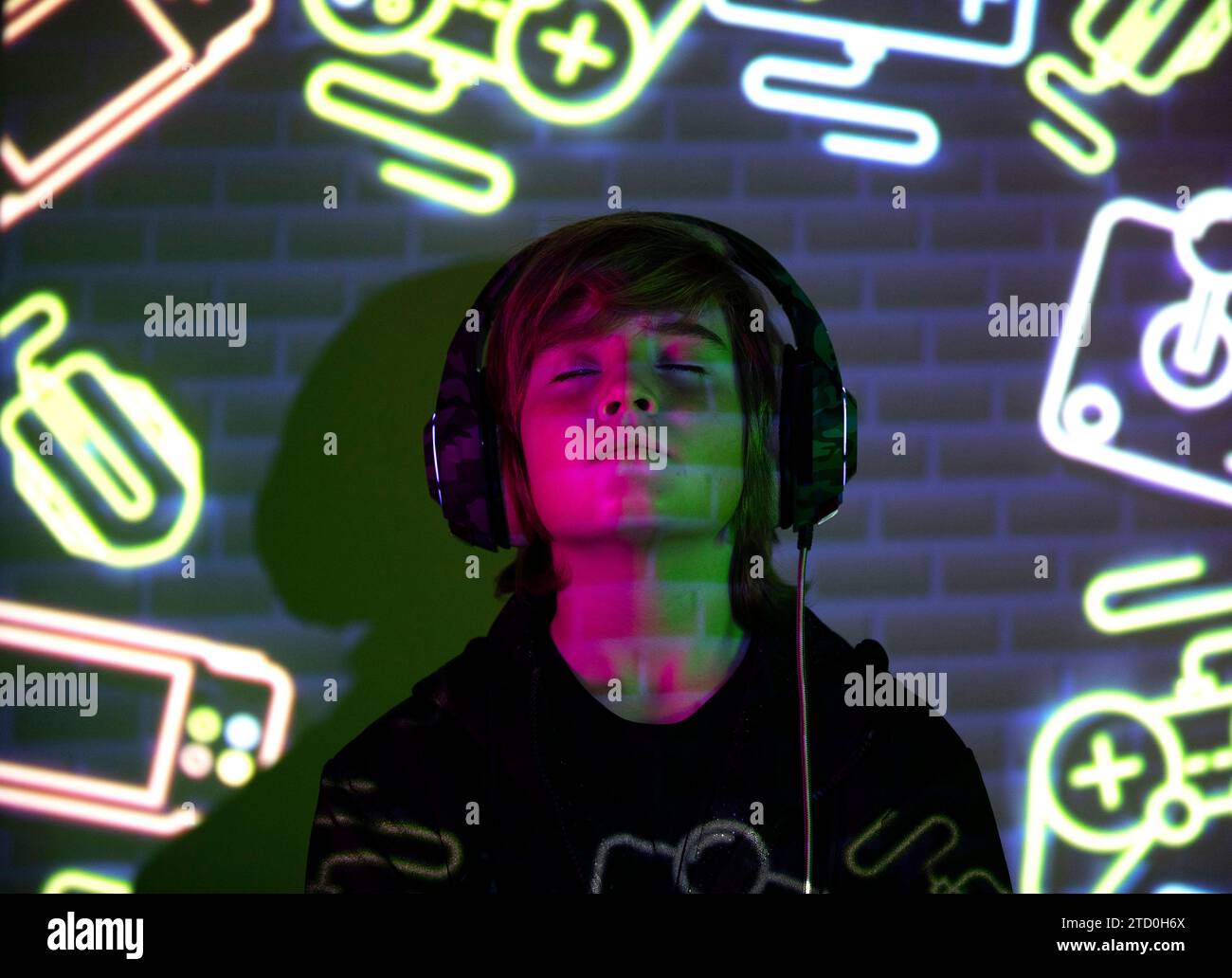 Ein junger Junge, der in der Musik verloren ist, mit farbenfrohen Neon-Gaming-Ikonen auf Gesicht und Hintergrund, die eine lebendige und moderne Atmosphäre schaffen. Stockfoto