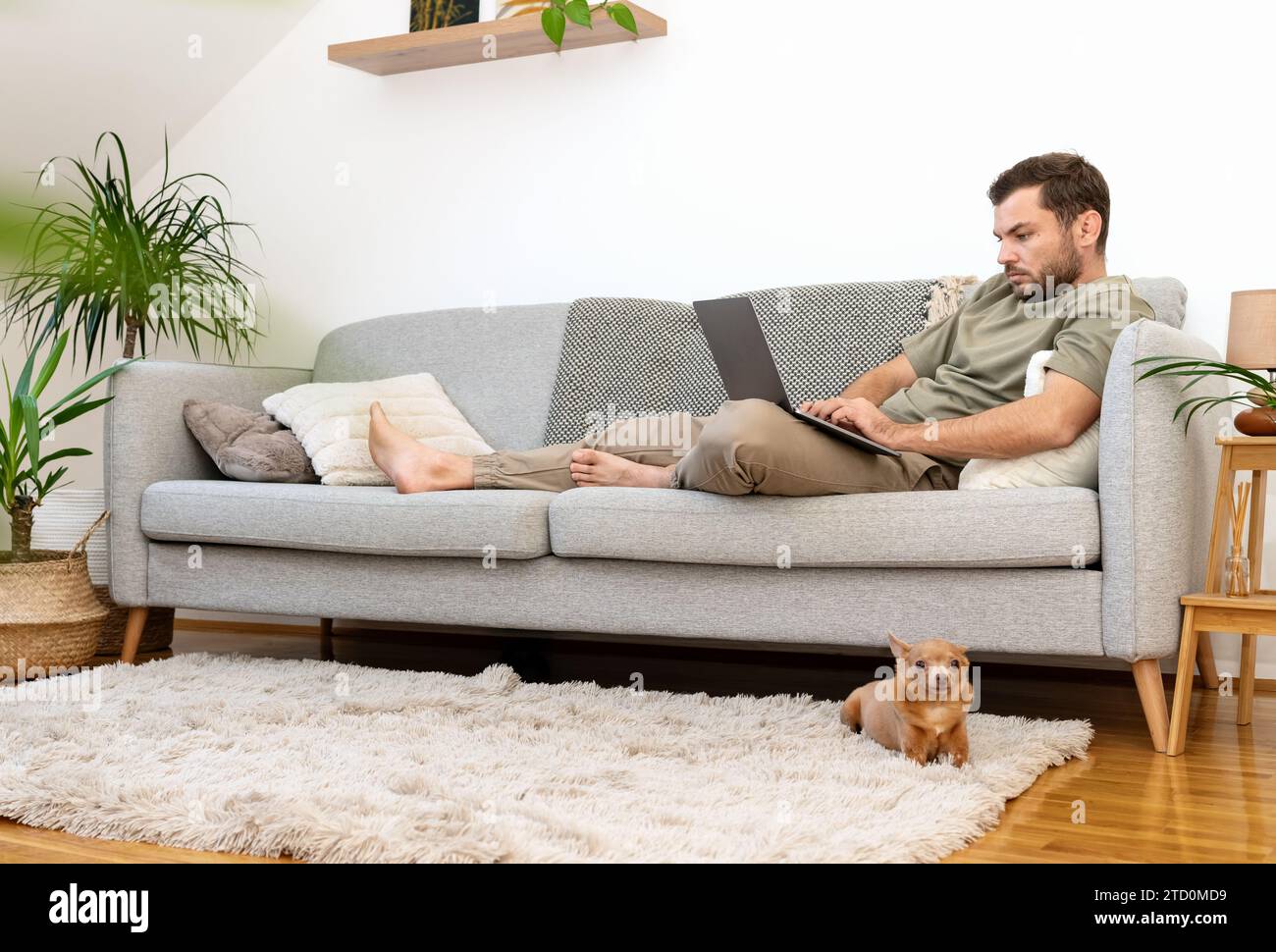Ein Mann, der sich zu Hause auf dem Sofa liegend und seinen Laptop benutzt. Arbeiten von zu Hause aus. Häuslicher Lebensstil. Stockfoto