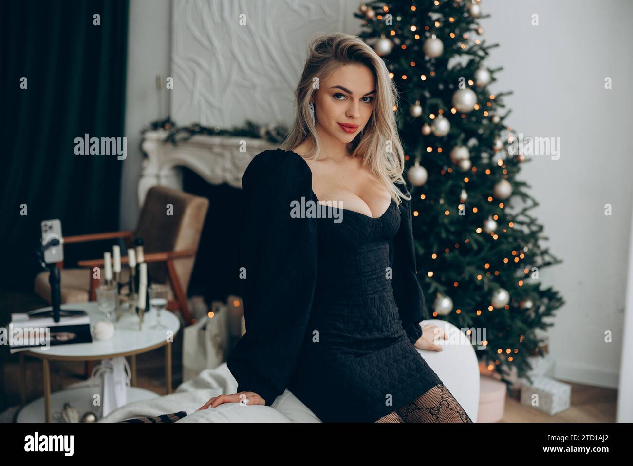 Junge verführerische blonde Frau, die neben einem geschmückten Weihnachtsbaum sitzt, in stilvollem schwarzen Kleid mit Dekolleté. Nahaufnahme. Stockfoto