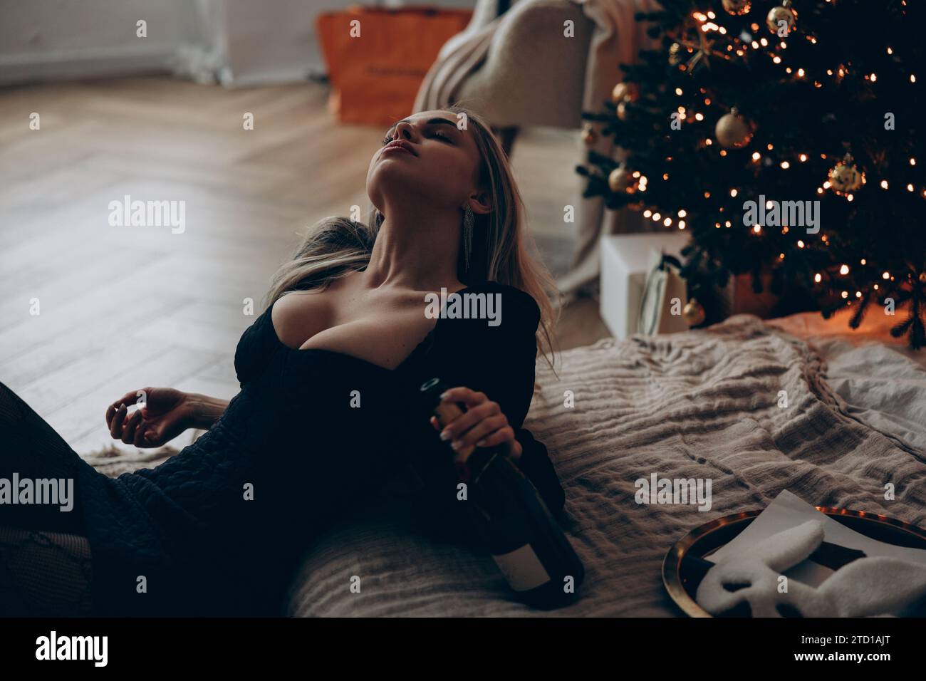 Junge blonde Frau, die neben dem Bett sitzt und sich in stilvollem schwarzen Minikleid mit Weinflasche in der Hand am Weihnachtsbaum entspannt. Nahaufnahme. Stockfoto