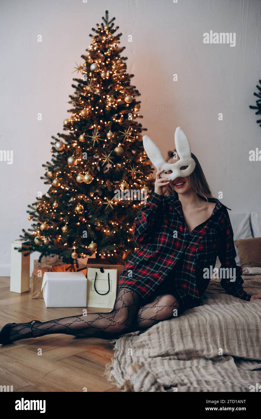 Junge verführerische Frau, die im karierten Hemd auf dem Bett sitzt und Hasenmaske im Gesicht vor dem Hintergrund des leuchtenden Weihnachtsbaums hält. Stockfoto