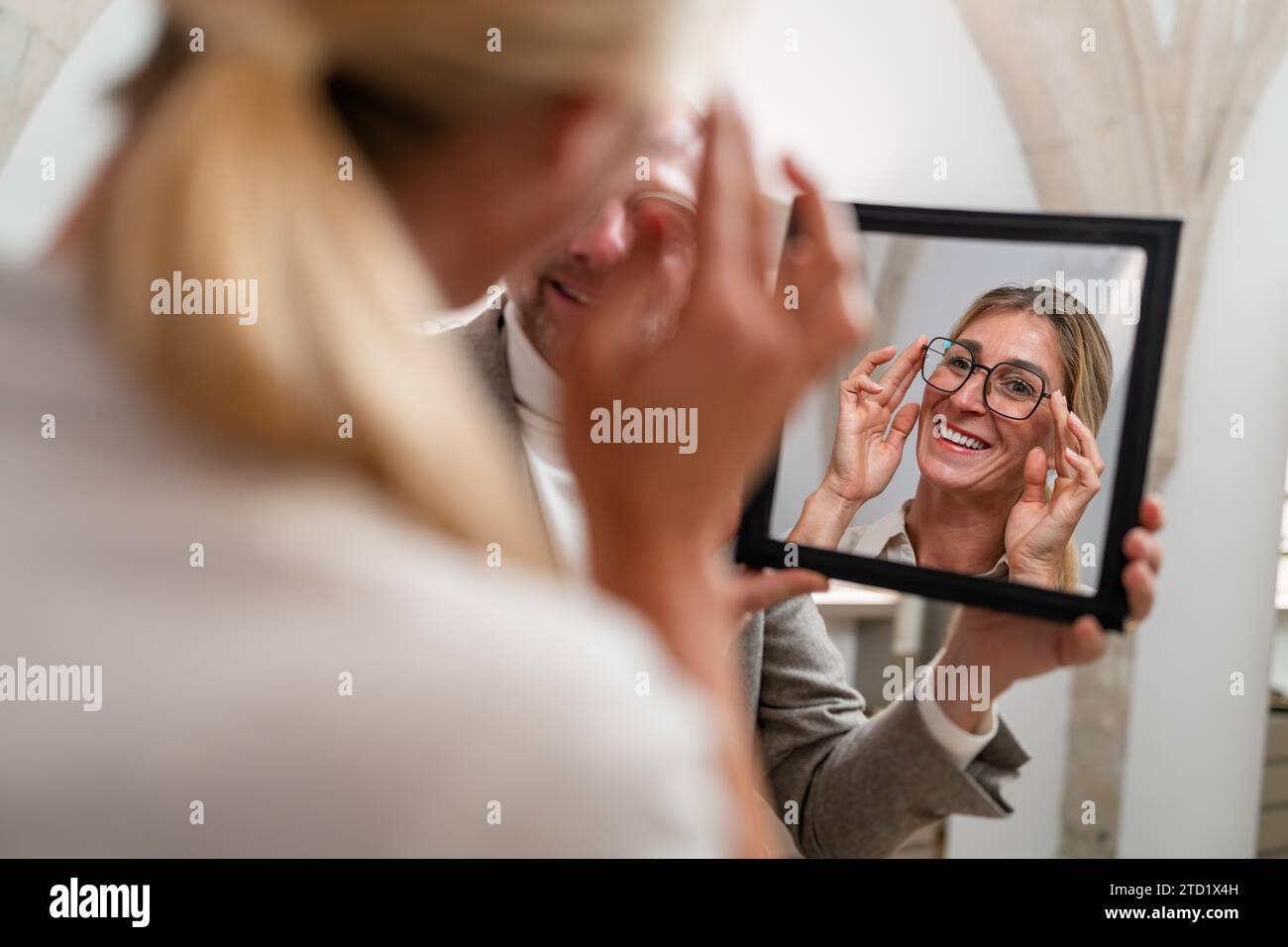 Optiker hält einen Spiegel vor einer lächelnden Frau, die in einem optischen Geschäft Brillen anprobiert. Stockfoto
