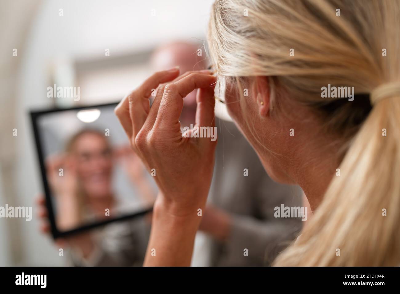 Frau, die eine Brille anprobiert und ihr Spiegelbild in einem Spiegel in einem Optikergeschäft ansieht. Der Fokus liegt auf Händen, die eine Brille mit verschwommenem Hintergrund halten Stockfoto