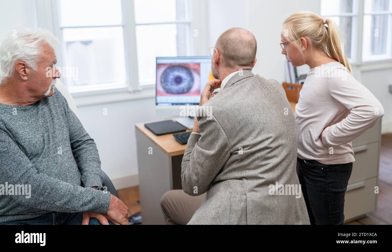Optometrist, der den Augenscan eines älteren Patienten auf einem Computer überprüft, sieht seine Enkelin aufgeregt zu. Stockfoto