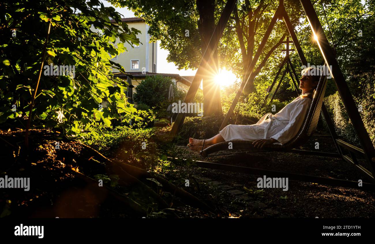 Frau in einem weißen Bademantel, die sich auf einer Korbschaukel im Garten des Wellnesshotels entspannt Stockfoto