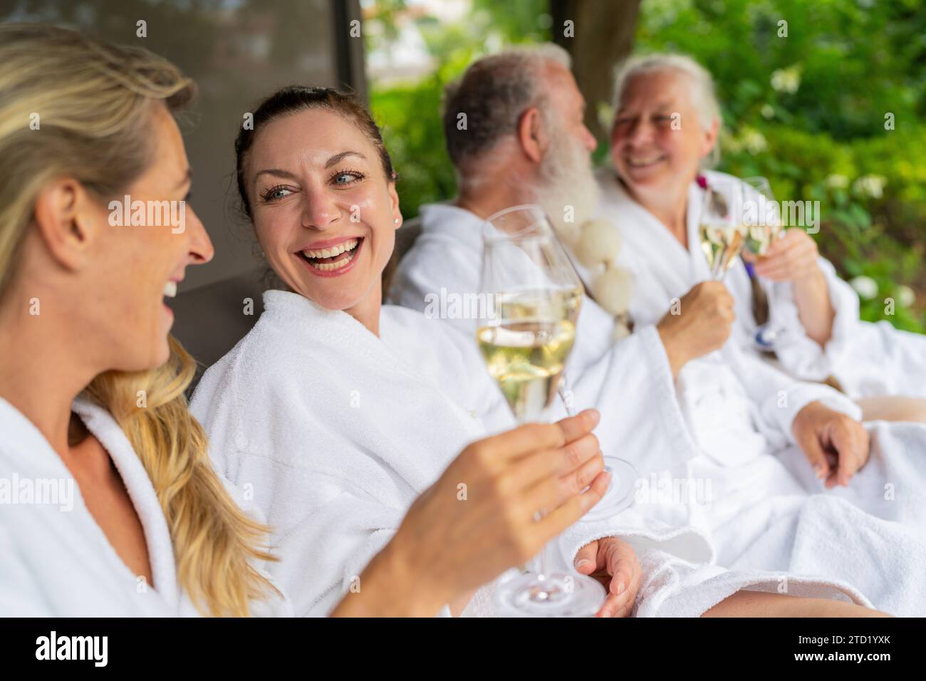 Gruppe von Menschen in weißen Bademänteln lachend und mit Sektgläsern in der Hand, die auf einer Liege in einem Wellnesshotel feiern Stockfoto