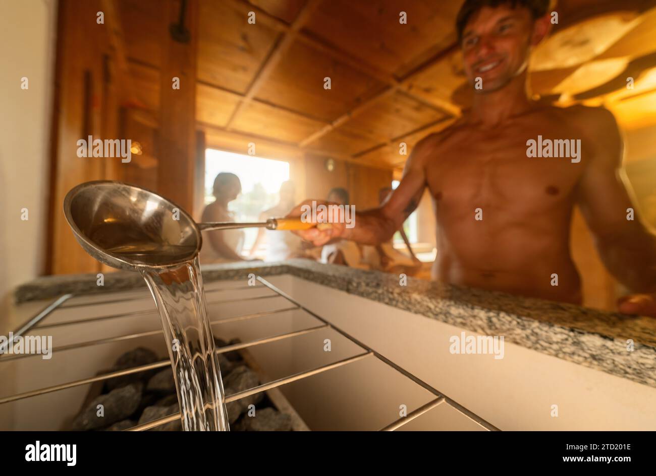 Nahaufnahme eines Mannes mit einer Kelle, der Wasser auf heiße Saunageleine gießt, mit einer verschwommenen Person im Hintergrund. Abschluss Sauna Spa Wellness Hotel Konzept Bild Stockfoto