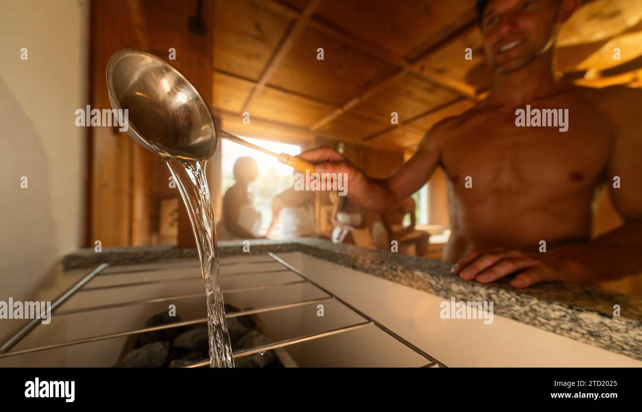 Mann gießt Wasser aus einer Saunakelle auf heiße Felsen auf einem Saunaofen. Menschen im Hintergrund. Abschluss Sauna Spa Wellness Hotel Konzept Bild. Stockfoto