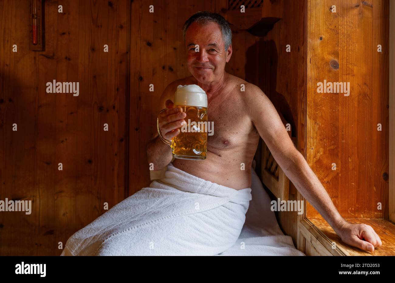 Ein lächelnder Mann in der Sauna hält einen deutschen Bierbecher, ein Handtuch um die Taille gewickelt. Bild des Spa Wellness-Konzepts Stockfoto