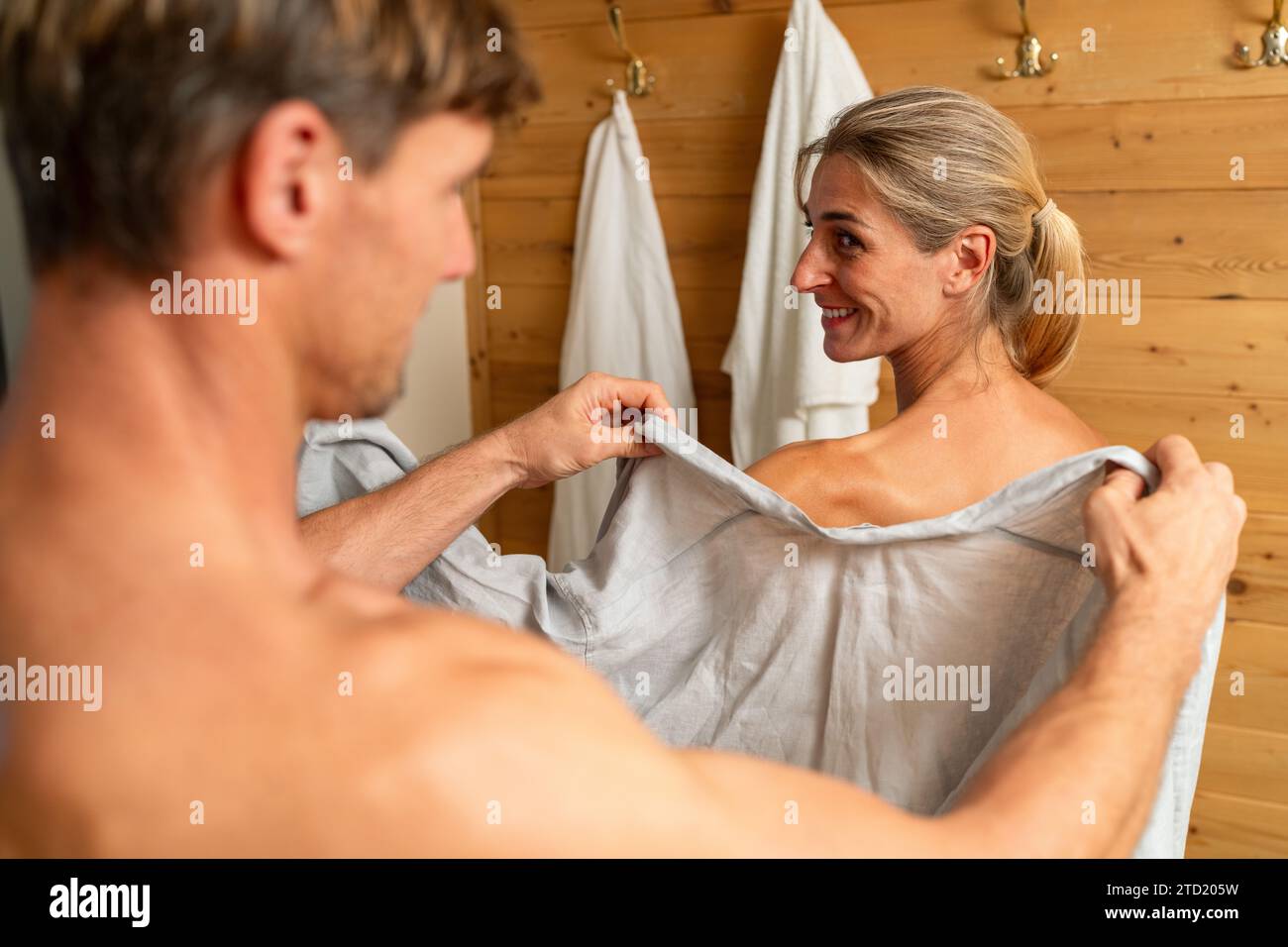 Mann, der einer Frau nach der Sauna einen Bademantel in einem Wellnessresort anbietet, beide ein Lächeln teilen Stockfoto