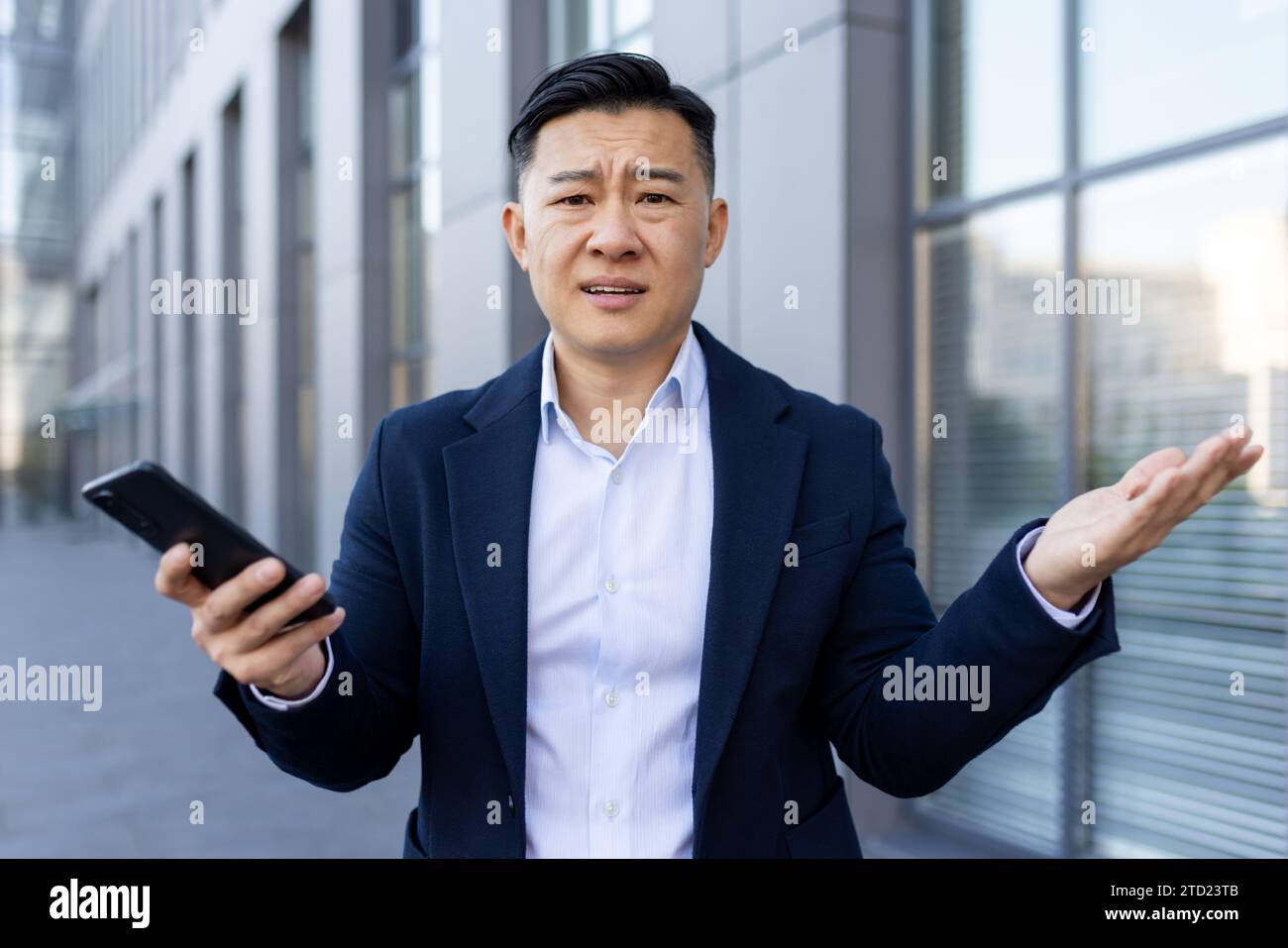 Porträt eines jungen asiatischen Geschäftsmannes, der in der Nähe eines Bürogebäudes steht und aufgebracht ist, seine Hände spreizt, das Telefon hält und wor aussieht Stockfoto