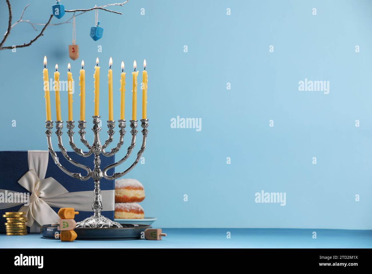 Hanukkah-Feier. Menora mit brennenden Kerzen, Dreidels, Donuts und Geschenkbox auf dem Tisch vor hellblauem Hintergrund. Leerzeichen für Text Stockfoto