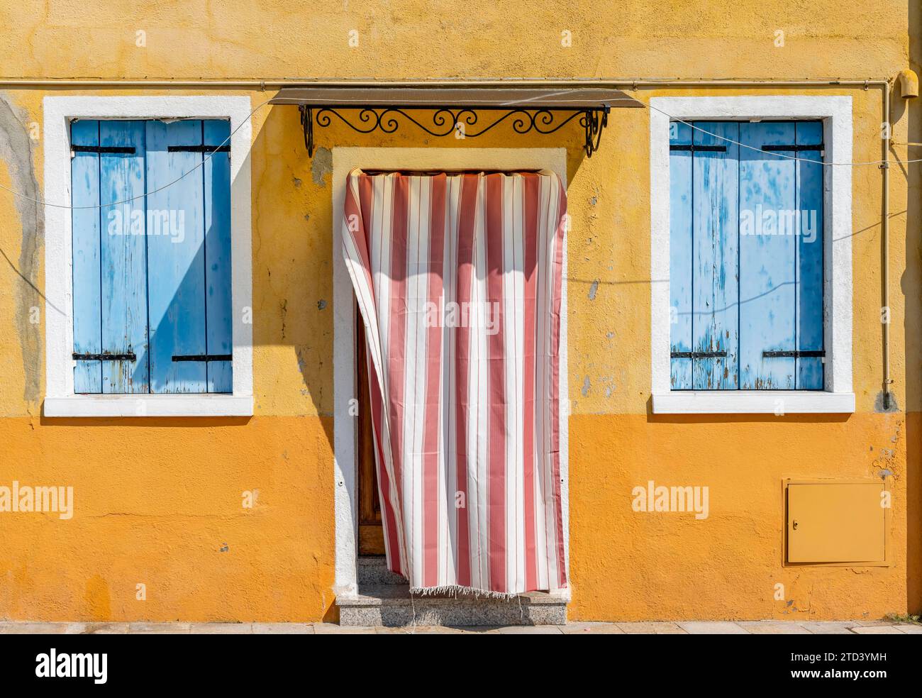 Gelbe Hausfassade mit Eingangstür mit rot-weißem Vorhang und Fenstern mit blauen Fensterläden, farbenfrohe Häuser auf der Insel Burano, Venedig Stockfoto