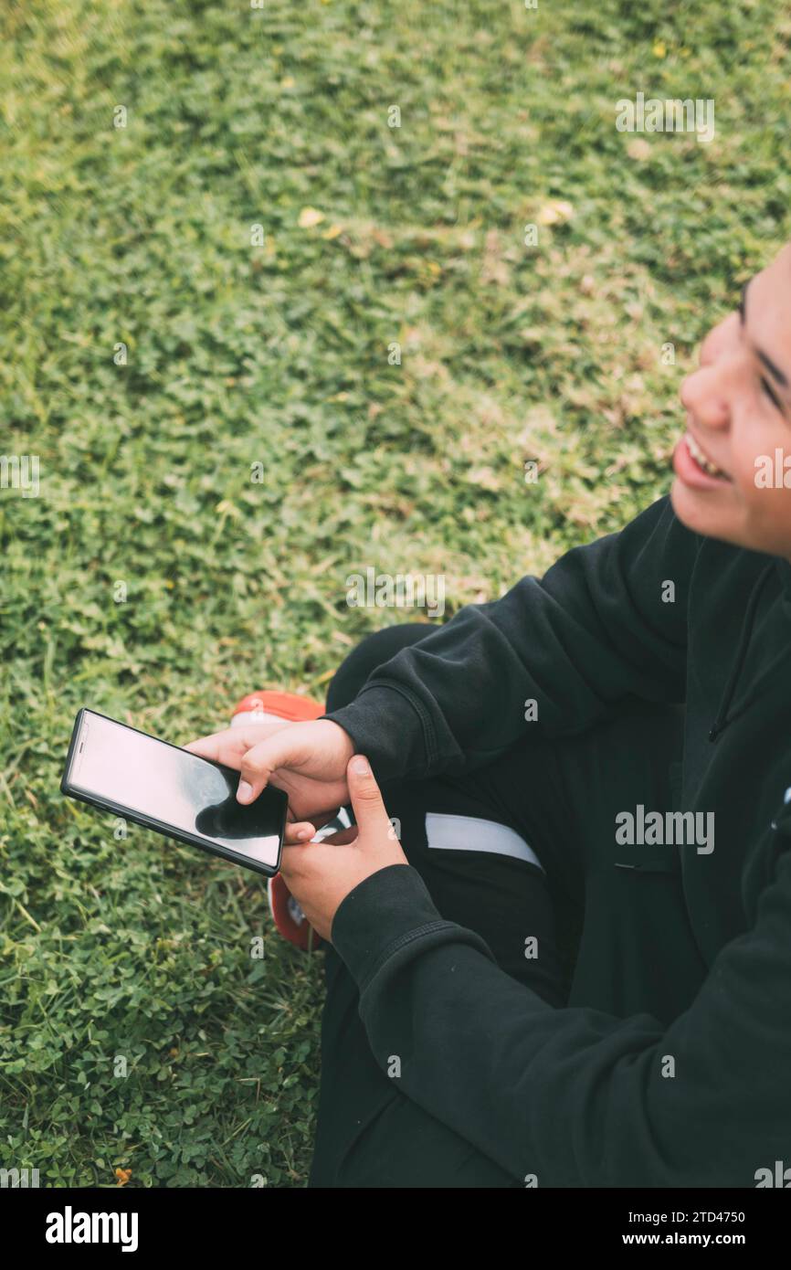 Ein fröhlicher Teenager mit Latino-Abstieg lächelt, während er auf seinen Smartphone-Bildschirm schaut. Die entspannte Haltung des Jungen und sein fröhlicher Ausdruck vermitteln ein Gefühl für Stockfoto