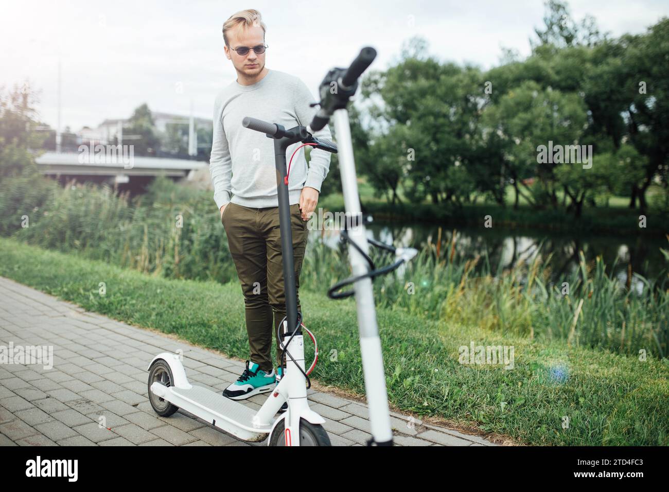 Der junge, gutaussehende blonde Mann wartet auf einen Freund, der mit Elektrorollern unterwegs ist Stockfoto