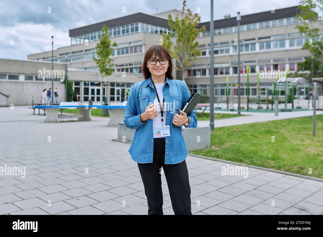 Schullehrerin mittleren Alters mit digitalem Tablet posiert in der Nähe des Schulgebäudes Stockfoto