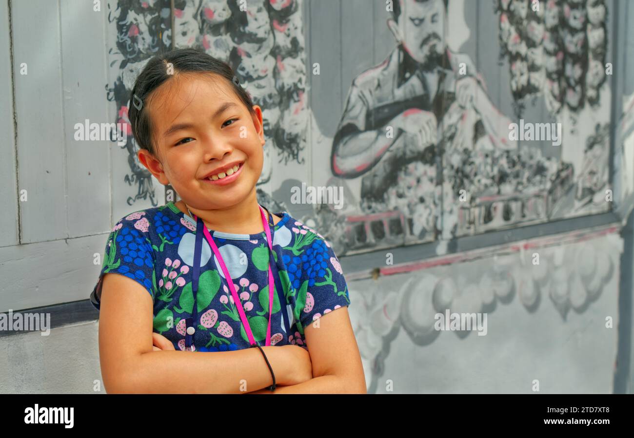Porträt eines asiatischen Mädchens im Alter von 9 oder 10 Jahren auf der Straße Thailands, Arme auf der Brust gekreuzt, lächelndes Gesicht, verschwommener Hintergrund eines Unident Stockfoto