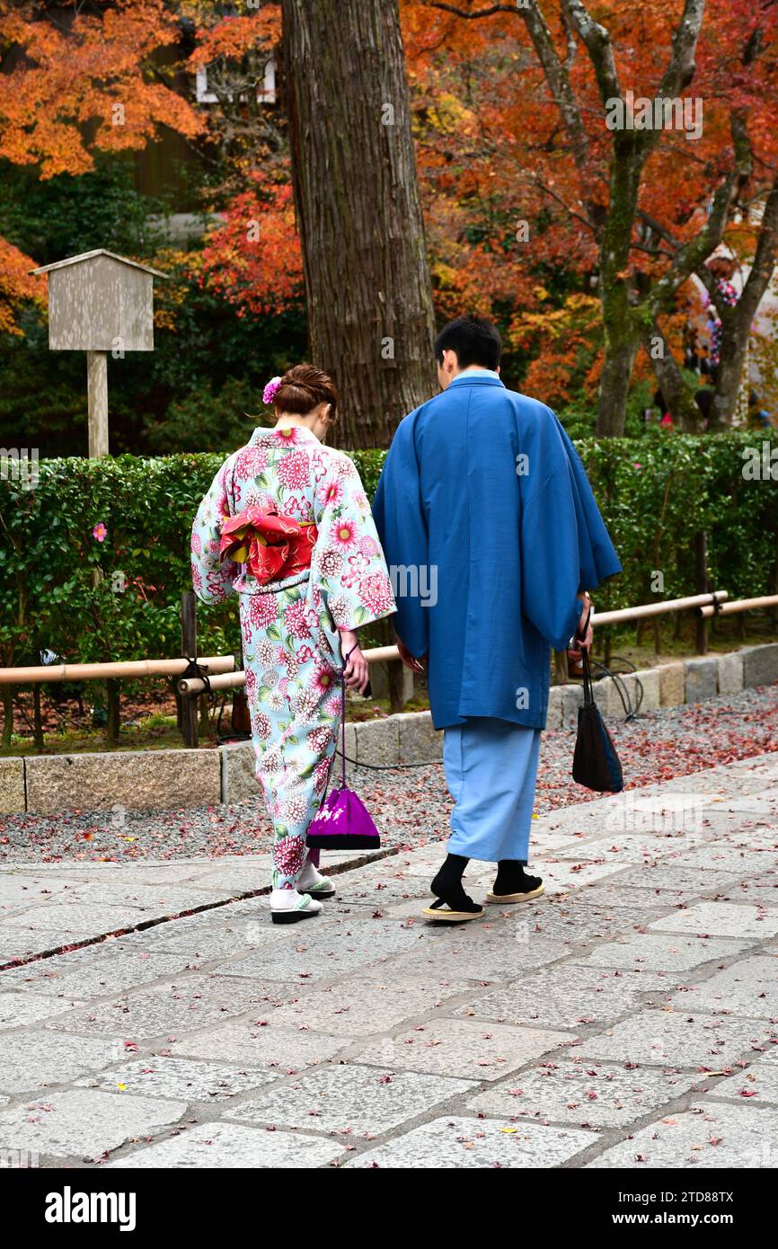 Japanisches Paar in traditioneller Kleidung, das auf dem Gehweg mit herbstlichen Blättern im Hintergrund spaziert. Stockfoto