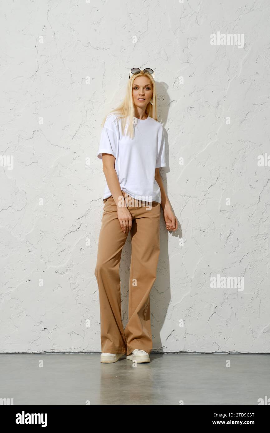 Trendige Erwachsene Frau in brauner, breiter Hose und weißem T-Shirt, das sich an der Studiowand anlehnt Stockfoto