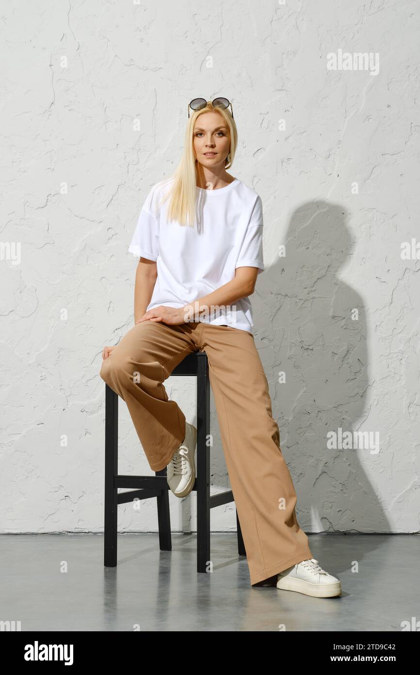 Trendige Erwachsene Frau im urbanen Outfit posiert auf hohem Hocker im weißen Studio Stockfoto