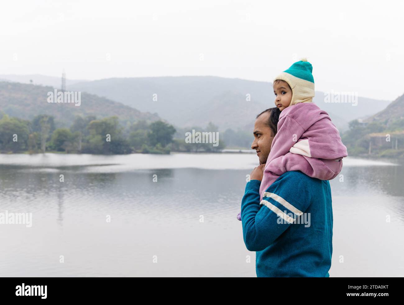Niedlicher Kleinkind Sohn, der auf der Schulter des jungen Vaters sitzt, im Wintertuch und in der Bergsee-Landschaft am Tag Bild wird im Jagdish-Tempel udaipur rajasthan aufgenommen Stockfoto