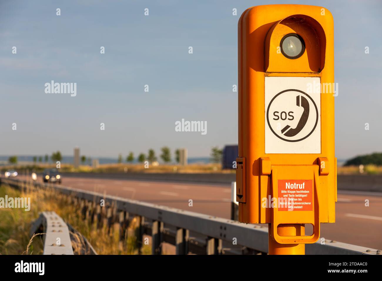 Straßenverkehrssicherheit: Oranges SOS-Anruffeld auf deutscher Autobahn unter verschwommenen Autolichtern, deutsche Textübersetzung: SOS, Notruf, bitte heben Sie die Klappe an Stockfoto