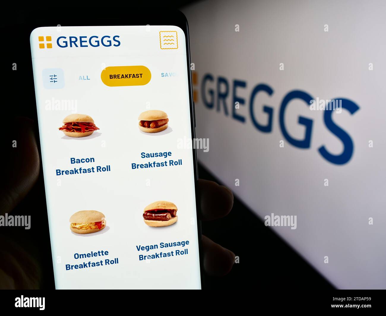 Person mit Smartphone und Webseite der britischen Bäckereikette Greggs plc vor dem Geschäftslogo. Konzentrieren Sie sich auf die Mitte des Telefondisplays. Stockfoto