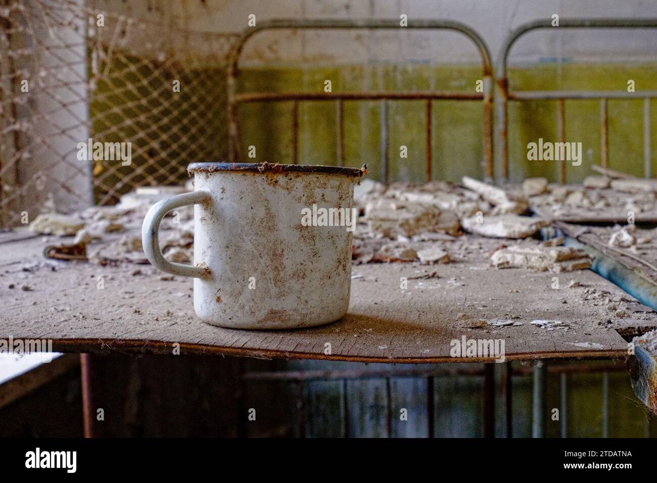 Eine verrostete Tasse auf einem baufälligen Tisch in einem verlassenen Zimmer. Ein weißer Metallbecher auf einem Bett in einem verlassenen Gebäude in Tschernobyl. Stockfoto