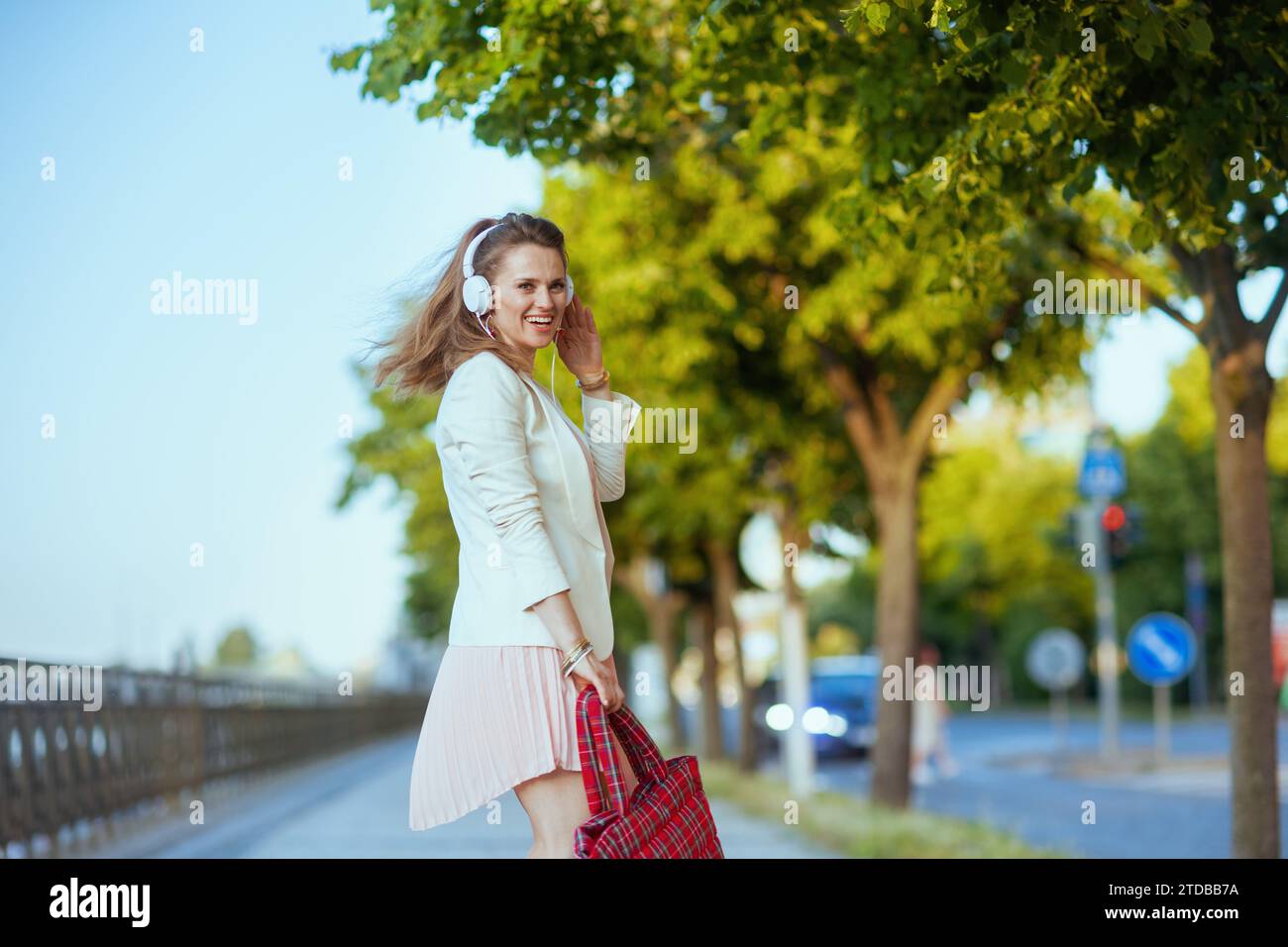 Lächelnde, elegante Frau in rosa Kleid und weißer Jacke in der Stadt, die Musik mit Kopfhörern hört. Stockfoto