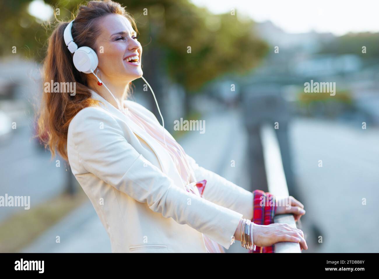 Lächelnde junge Frau in rosa Kleid und weißer Jacke in der Stadt mit Kopfhörern. Stockfoto