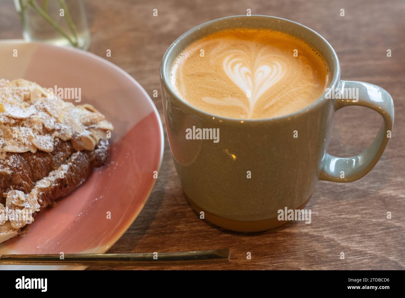 Ein Latte in einer Kaffeetasse, der morgens in einem Café neben einem Mandelcroissant getrunken wird. Herzsymbol in der Creme auf dem Kaffee. UK Stockfoto