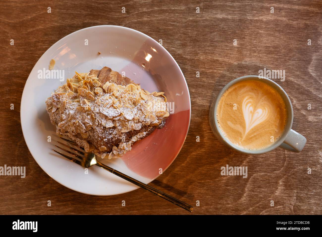 Ein Latte in einer Kaffeetasse, der morgens in einem Café neben einem Mandelcroissant getrunken wird. Herzsymbol in der Creme auf dem Kaffee. UK Stockfoto