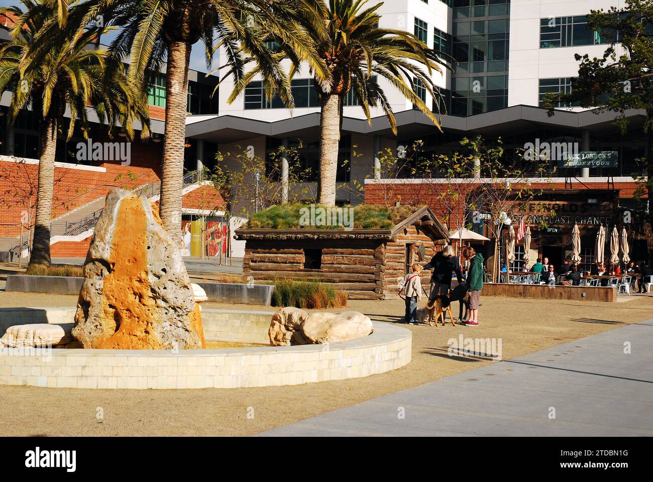 Eine Familie spaziert mit ihrem Hund auf dem Jack London Square, in der Nähe einer Nachbildung der Blockhütte des Autors in Call of the Wild in Oakland, Kalifornien Stockfoto