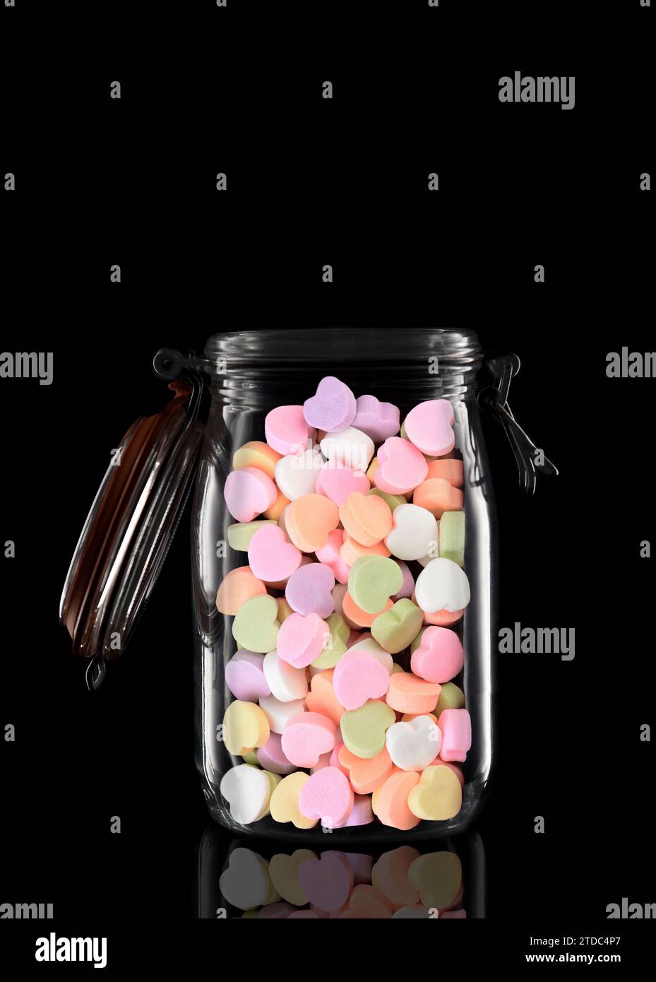 Ein Glasvorratsbehälter oder Dosenbecher gefüllt mit Valentines Candy Hearts isoliert auf Schwarz mit Reflexion, mit offenem Deckel. Stockfoto