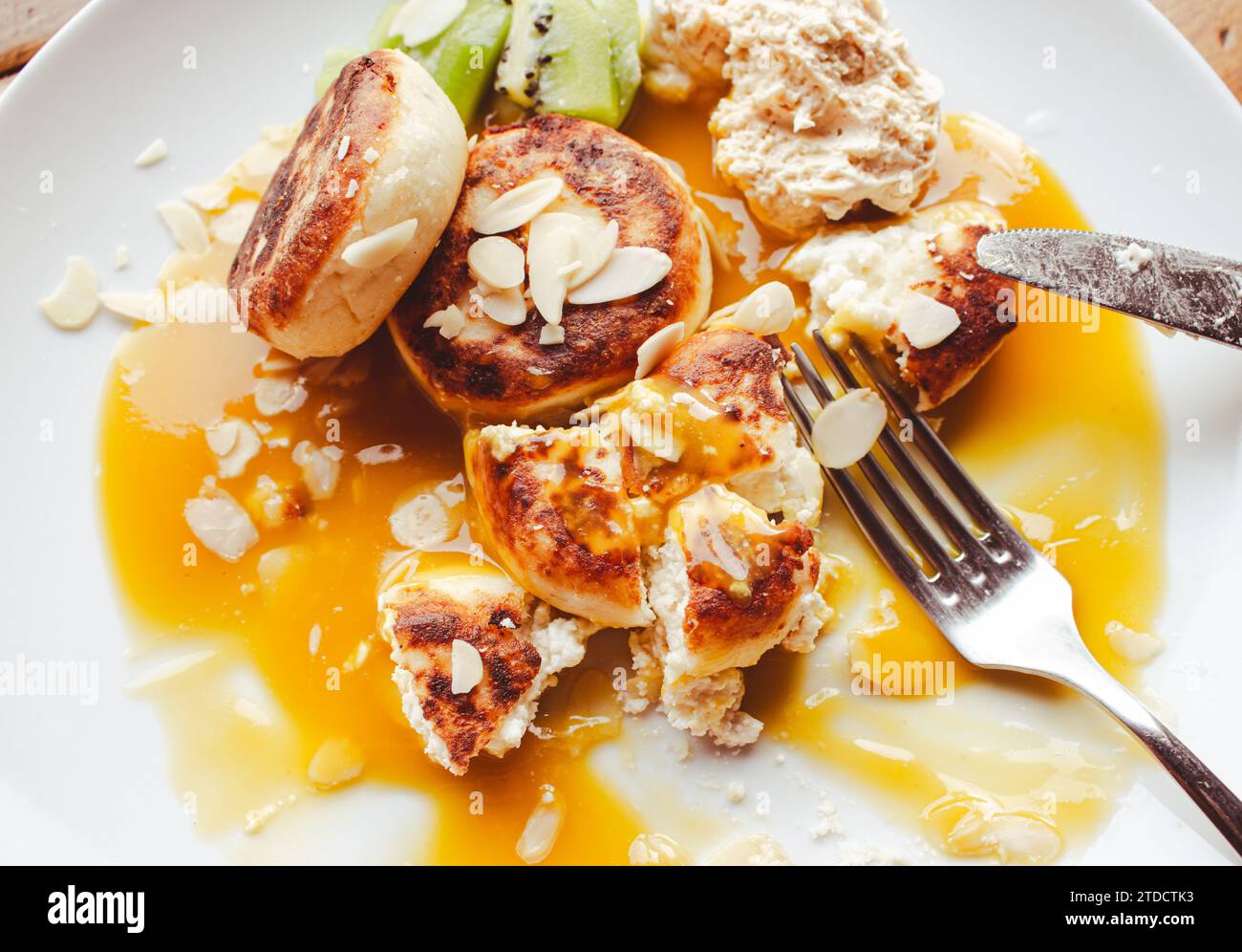 Käsekuchen mit Mangohyrup und Mandelflocken auf weißem Teller. Süßes Frühstück im Café. Syrnik mit Hütte und Avocado. Käsepfannkuchen mit Sirup. Stockfoto