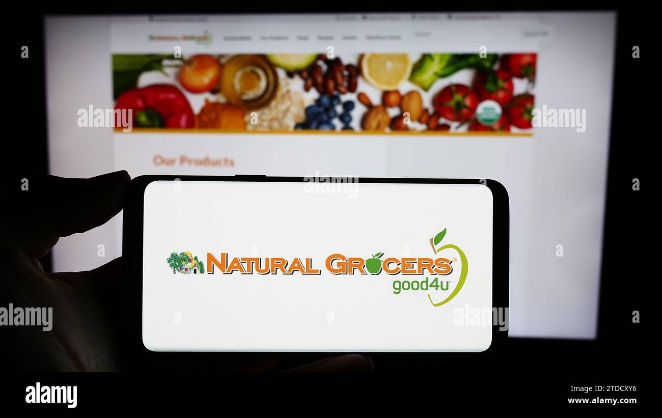 Person, die ein Mobiltelefon mit dem Logo des Einzelhandelsunternehmens Natural Grocers von Vitamin Cottage Inc. Vor der Webseite hält. Schwerpunkt auf dem Display des Telefons. Stockfoto
