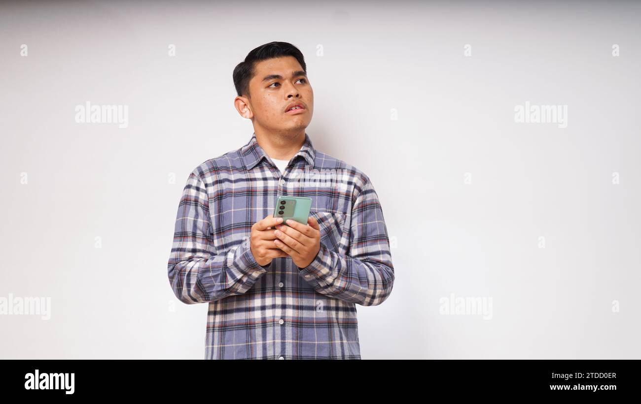 Junger asiatischer Mann, der sein Handy mit traurigem und verwirrtem Ausdruck auf weißem Hintergrund hält. studio-Aufnahme Stockfoto