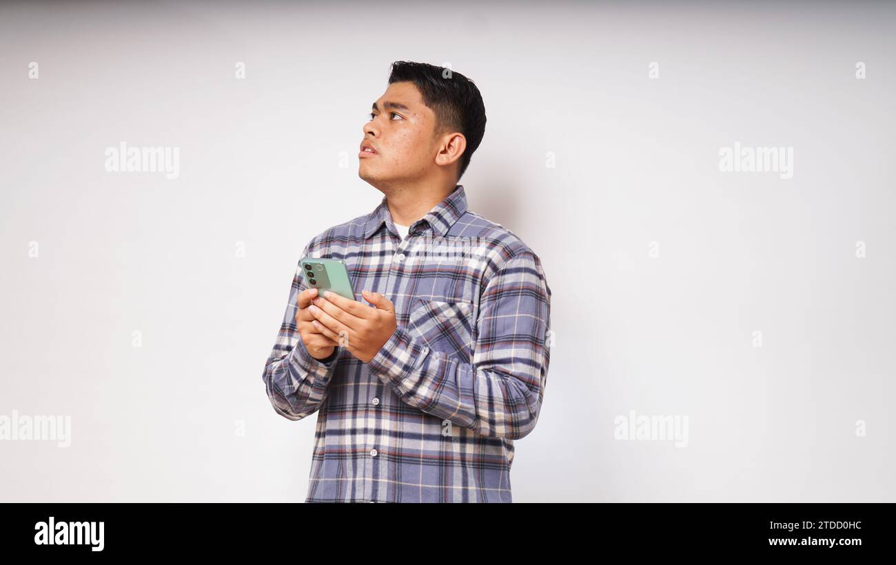 Junger asiatischer Mann, der sein Handy mit traurigem und verwirrtem Ausdruck auf weißem Hintergrund hält. studio-Aufnahme Stockfoto