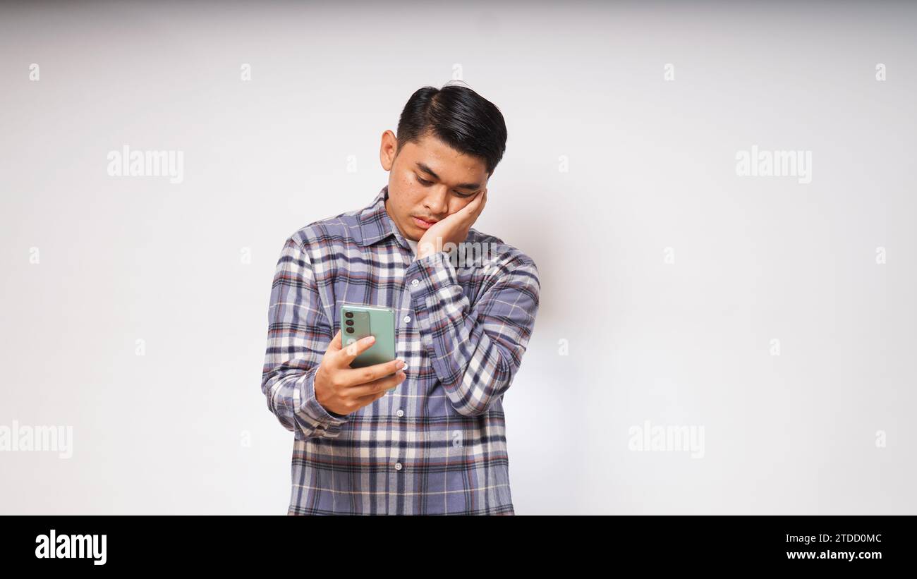 Junger asiatischer Mann, der sein Handy mit traurigem und verwirrtem Ausdruck auf weißem Hintergrund hält. studio-Aufnahme Stockfoto Junger asiatischer Mann, der sein Handy mit traurigem und verwirrtem Ausdruck auf weißem Hintergrund hält. studio-Aufnahme Stockfoto