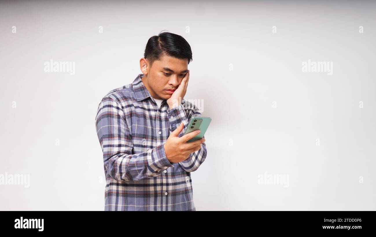 Junger asiatischer Mann, der sein Handy mit traurigem und verwirrtem Ausdruck auf weißem Hintergrund hält. studio-Aufnahme Stockfoto Junger asiatischer Mann, der sein Handy mit traurigem und verwirrtem Ausdruck auf weißem Hintergrund hält. studio-Aufnahme Stockfoto