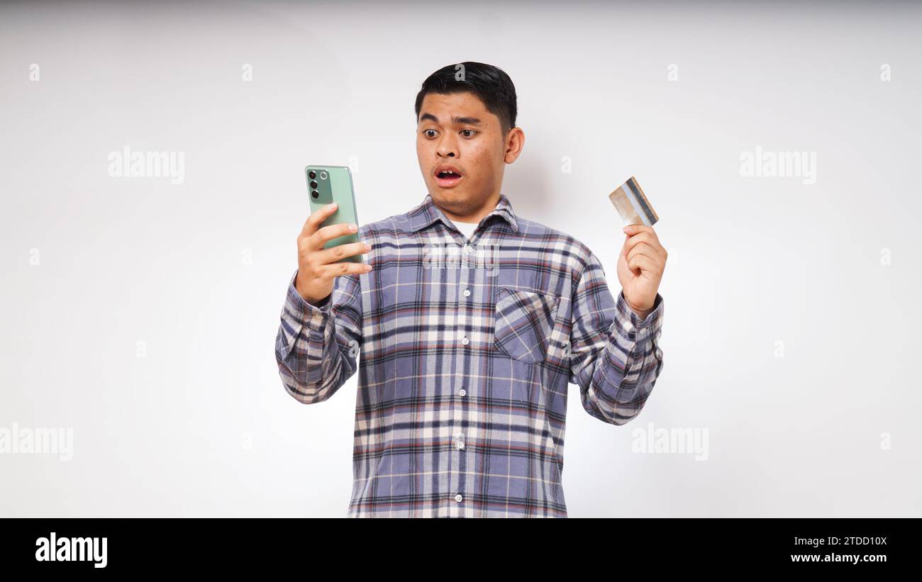 Asiatischer junger Mann mit schockiertem Ausdruck, der Kreditkarte und Smartphone hält, mit großen Augen auf den Smartphone-Bildschirm blickt, Innenstudio-Aufnahme isoliert Stockfoto