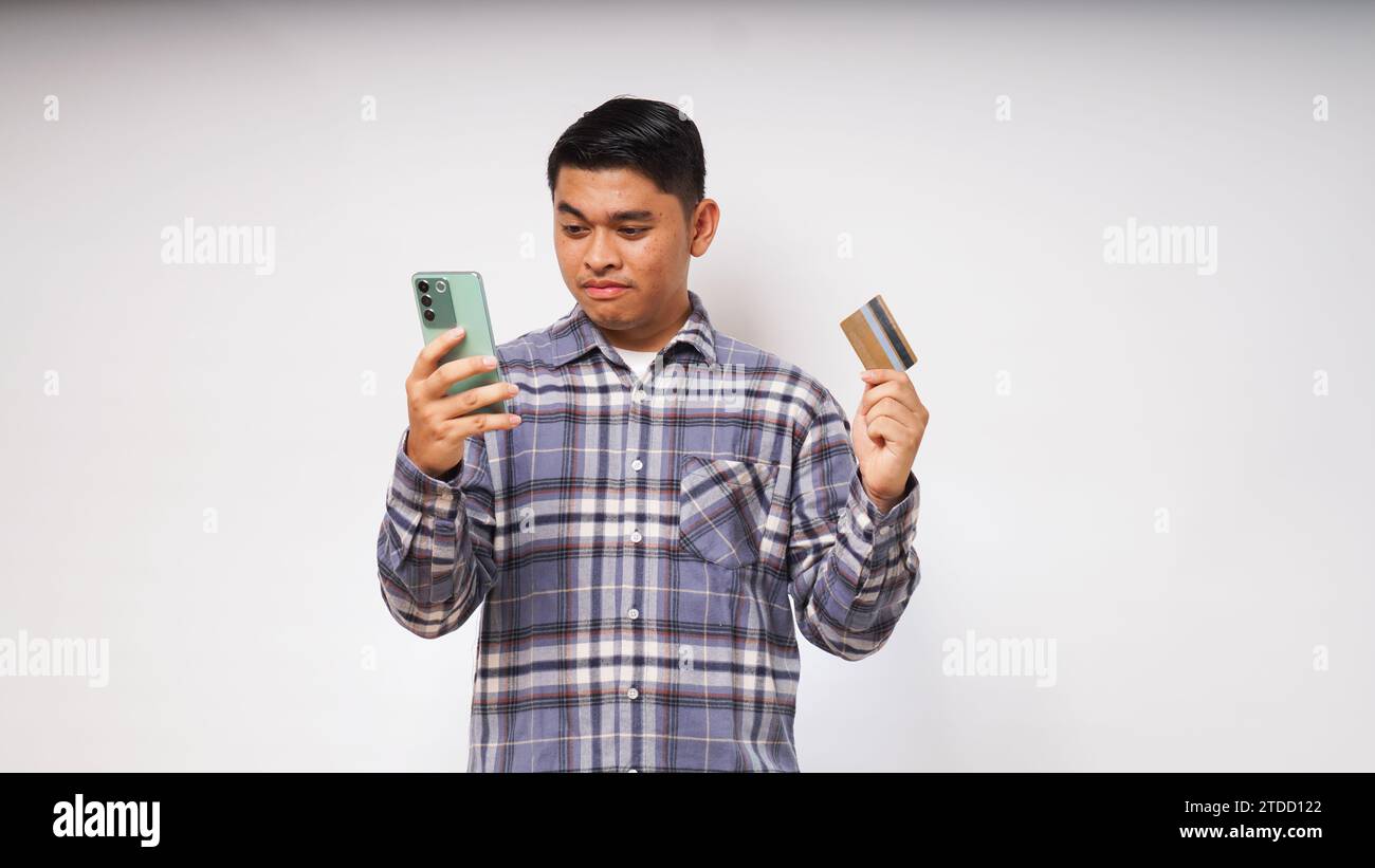 Asiatischer junger Mann mit schockiertem Ausdruck, der Kreditkarte und Smartphone hält, mit großen Augen auf den Smartphone-Bildschirm blickt, Innenstudio-Aufnahme isoliert Stockfoto